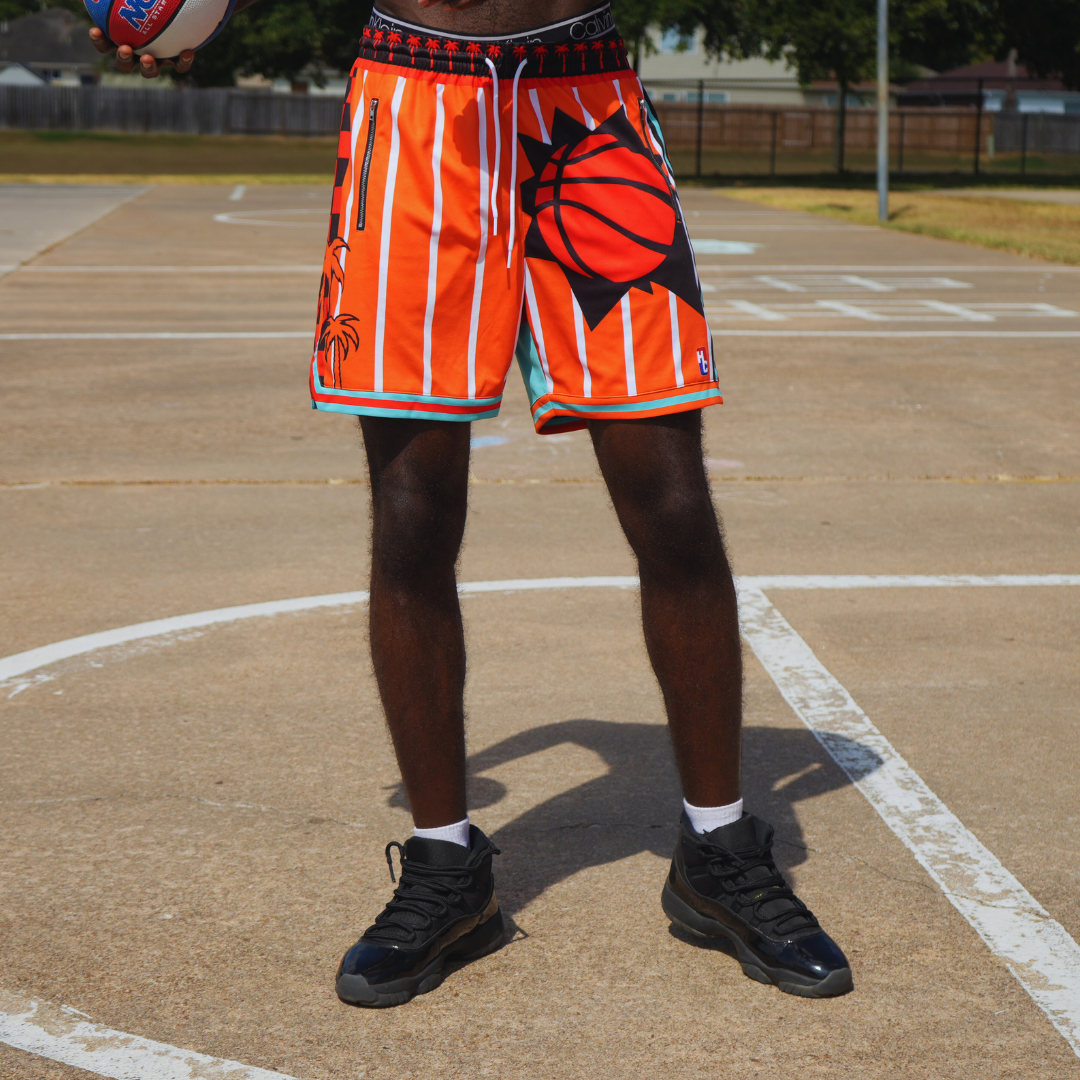Tropical Heat Shorts in vibrant orange with black and white basketball design, mesh fabric, zipper pockets, and drawstrings on a basketball court.