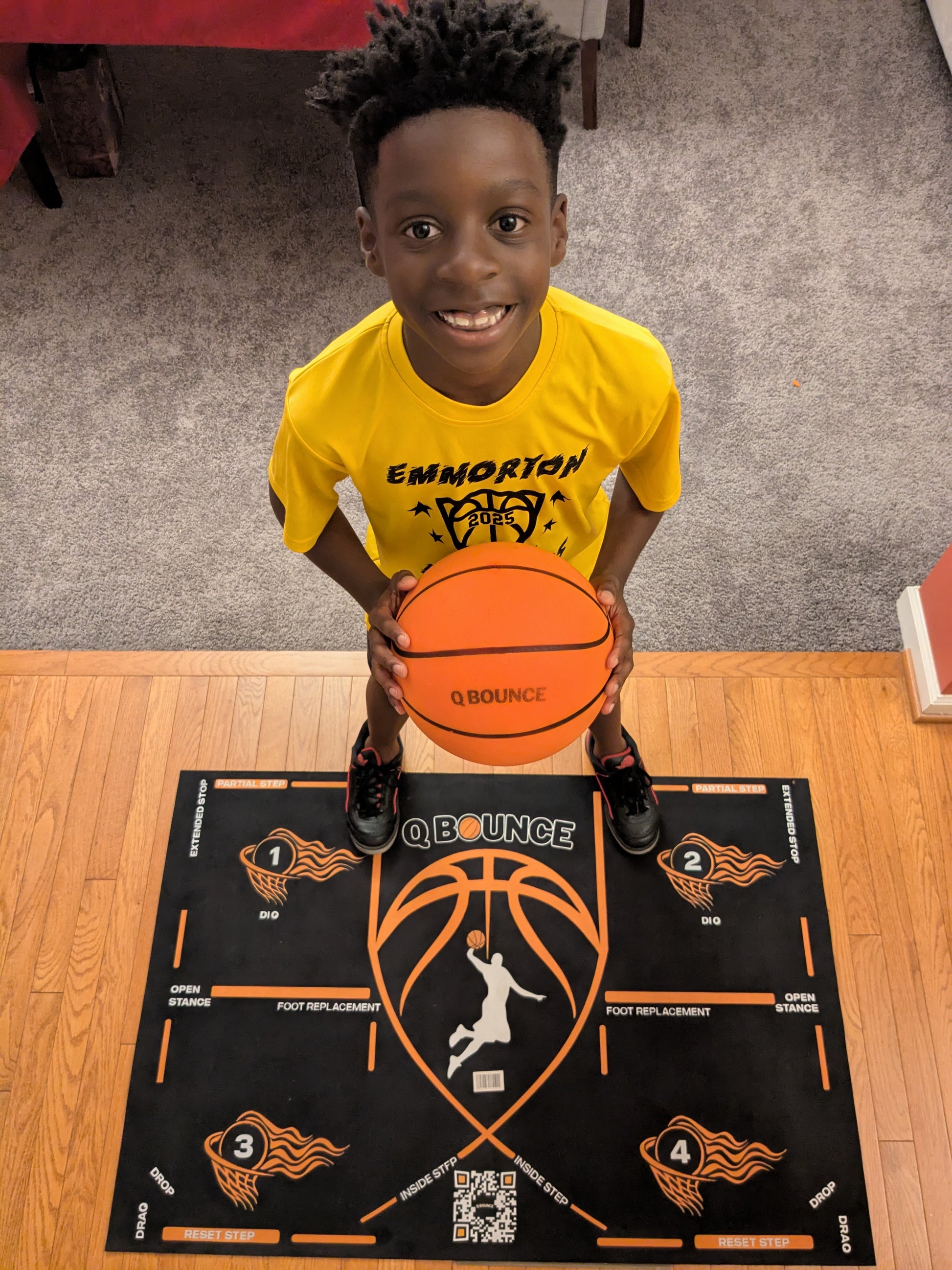 Boy in yellow shirt holding basketball on QBounce Basketball Starter Bundle training mat indoors