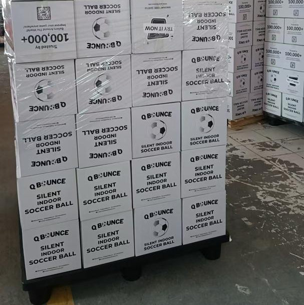 Stacked boxes of QBounce Silent Football 2.0 packaging ready for shipment in a warehouse setting.